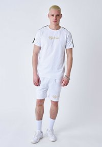 Conjunto de camiseta blanca y pantalones cortos de algodón, con branding dorado en el pecho y en la pierna, y detalles laterales en negro. Incluye zapatillas blancas y calcetines.