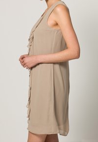 Beżowa sukienka bez rękawów wykonana z lekkiego materiału, z falbaniastym detalem z przodu i luźnym krojem. Kończy się powyżej kolana.