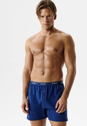 Młody mężczyzna z krótkimi brązowymi włosami w niebieskich bokserkach marki Atlantic stojący na tle jednolitego jasnego tła.