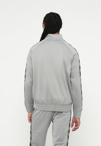 Persona con el cabello trenzado largo, vestida con un chándal gris claro con rayas de patrones negros y blancos en las mangas y piernas del pantalón, de espaldas.