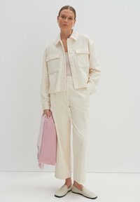 Cremfarbene Cordjacke mit zwei Brusttaschen, passenden Hosen und einem hellrosa Schal. Das Model trägt beigefarbene Slipper und steht vor einem weißen Hintergrund.
