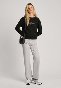 Sudadera negra con logo gráfico, jeans de pierna ancha gris y zapatillas plateadas. Se lleva un pequeño bolso de mano negro. El cabello lacio cae sobre los hombros.