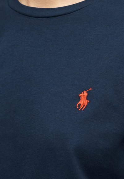 Κοντινή λήψη μπλε υφάσματος ναυτικού χρώματος με μικρό πορτοκαλί κεντημένο λογότυπο παίκτη πόλο πάνω σε άλογο που κρατά ανυψωμένη ρακέτα.