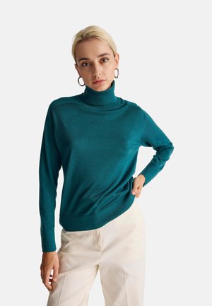 Türkiser Rollkragenpullover mit weicher Textur und figurschmeichelndem Design, ausgestattet mit langen Ärmeln und einer lockeren Passform an der Taille. Kombiniert mit weißen Hosen.