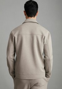 Sweatshirt bej cu guler, textură netedă și detalii de cusătură. Prezentat din spate, împreună cu pantaloni asortați, într-un decor minimalist.