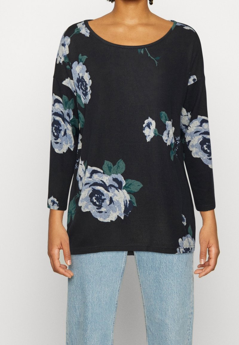 Svart långärmad topp med stora blå och gröna blommönster, rund halsringning och avslappnad passform, kombinerad med ljusblå jeans.