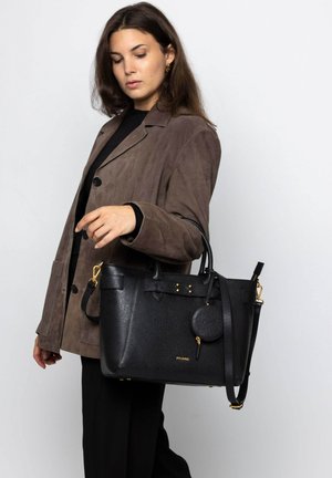 Zwarte leren schoudertas met een textuur oppervlak, gouden accenten en een ronde label. Model draagt een bruine suède jas, met een eenvoudige zwarte top.