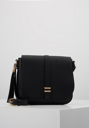 Black pebbled leather crossbody bag with front flap, gold buckle detail, adjustable strap, and side tassel charm on a white surface.