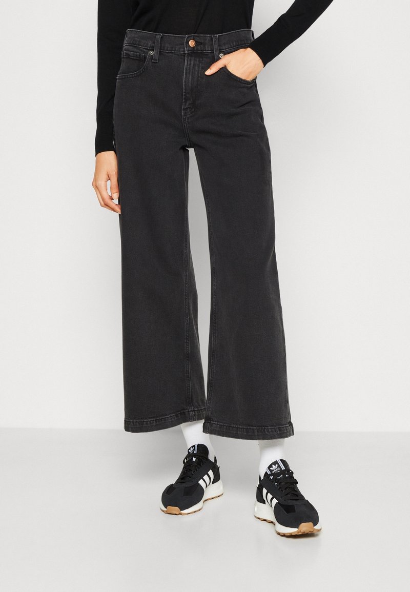 Jeans noirs à jambes larges avec une taille haute, dotés d'une fermeture à bouton à l'avant et de poches latérales, associés à des baskets noires et des chaussettes blanches.