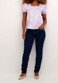Donna che indossa un top corto a maniche corte di tessuto lilla, jeans skinny blu scuro e décolleté beige a punta, in piedi davanti a un muro semplice.