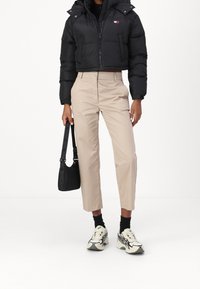 Persona con un giubbotto imbottito nero corto, pantaloni beige a vita alta, calzini neri, sneakers bianche e che tiene una borsa nera.