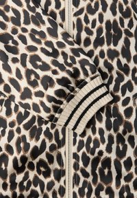 Ärmel einer Jacke mit Leopardenmuster und elastischem Bündchen mit schwarzen und beigen Streifen sowie sichtbarem silberfarbenem Reißverschluss auf beigem Hintergrund.