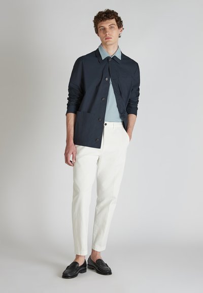 Granatowa kurtka zapinana na guziki z kieszenią na piersi, jasnobłękitna koszula pod spodem, białe dopasowane spodnie oraz czarne mokasyny. Gładka tekstura materiału.