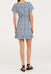 Robe à imprimé floral bleu avec des accents blancs, manches courtes volantées, taille plissée et ourlet à volants, associée à des sandales noires à brides.