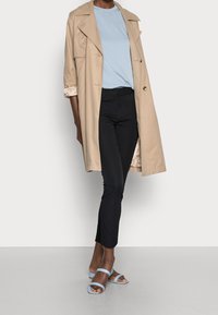 Trench-coat beige avec larges revers, t-shirt bleu clair, pantalon slim noir et sandales bleues à lanières. Textures lisses et coupe ajustée.