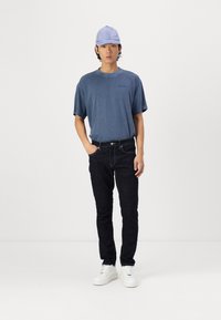 Blue relaxed-fit cotton t-shirt, black denim jeans, and white sneakers. Light blue cap with a slight sheen, standing against a plain backdrop.