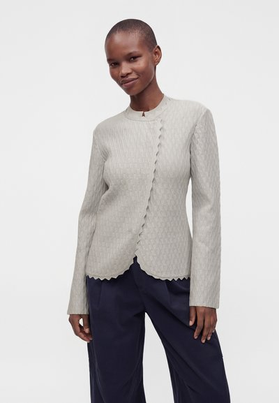 Personne debout et souriante, portant une veste gris clair texturée à bord festonné et un pantalon ample bleu marine foncé, sur un fond blanc uni.