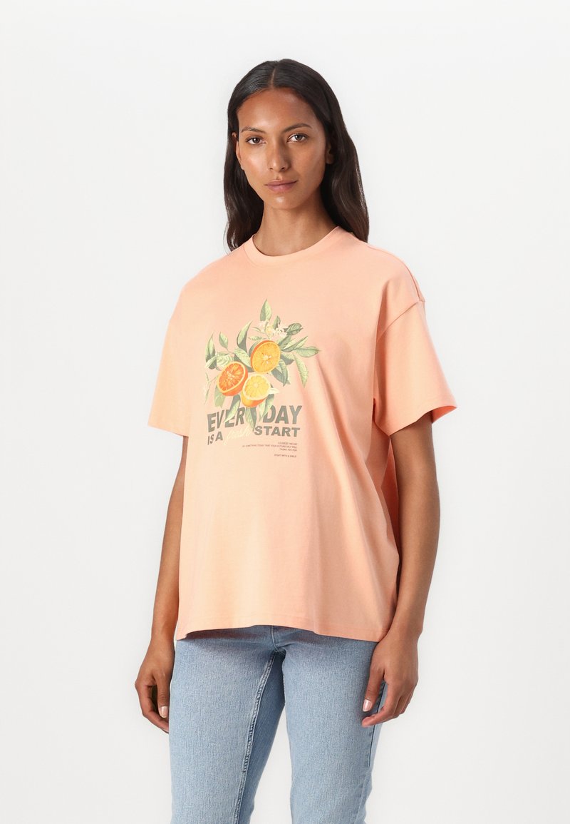 Camiseta de algodón melocotón de mangas cortas, con un gráfico floral de naranjas y hojas, y texto en verde y negro, con cuello redondo.