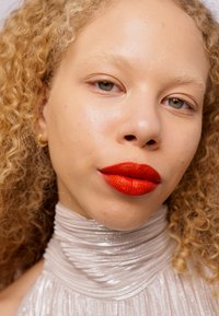 Lähikuva henkilöstä, jolla on kihara vaalea hius ja vaalea iho, ja hän pukeutuu hopeanväriseen teksturoituun korkeakauluksiseen puseroon sekä kirkkaan punaiseen huulipunaan.