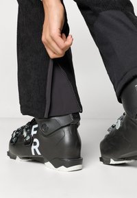 Μαύρο σορτς σκι με ραβδωτή υφή διαθέτει πλευρικό άνοιγμα με φερμουάρ. Ένα μαύρο μποτάκι σκι εμφανίζει λευκές σημάνσεις και ασημένιες αγκράφες.