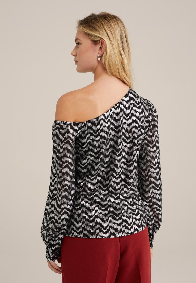 Schulterfreie Bluse aus transparenter schwarzer Stoff mit weißen Zickzackmustern, langen Puffärmeln und gerafftem Taillenbereich. Kombiniert mit roten Hosen.