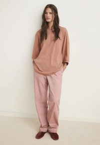 Overdreven rustkleurig top met losse mouwen, gecombineerd met lichtroze broek. Zachte stof, relaxte pasvorm en platte kastanjebruine schoenen.