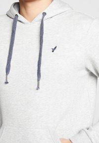 Grå hoodie med en strukturerad yta, utrustad med marinblå dragsko, känguruficka och en liten fågellogo broderad i marinblått på bröstet.