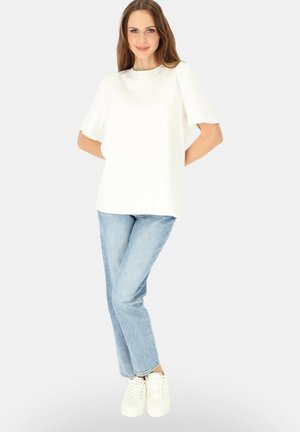 Kobieta stoi z rękami za plecami, ubrana w biały krótki top, jasnoniebieskie dżinsy i białe sportowe buty na jednolitym tle.