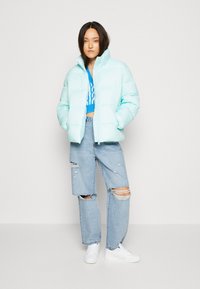 Lyse turkis pufferjakke med høy krage, oversized passform og quiltet design. Båret over en blå strikket crop top og slitte jeans.