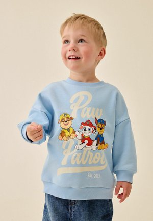 Junge lächelt und trägt einen hellblauen Pullover mit Paw Patrol Figuren und Text vorne, kombiniert mit blauen Jeans.