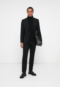 Completo nero con una vestibilità sartoriale, indossato su un dolcevita nero. Include una borsa in pelle nera. Le scarpe sono in pelle nera lucida.