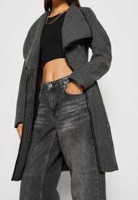 Harmaa villasekoitetakki, jossa on ylisuuri kaulus ja vetoketjuyksityiskohtia, yhdistetty tummanharmaisiin farkkuihin ja mustaan, ribbineulottuun lyhytpaitaan. Ketjusomiste näkyvissä.