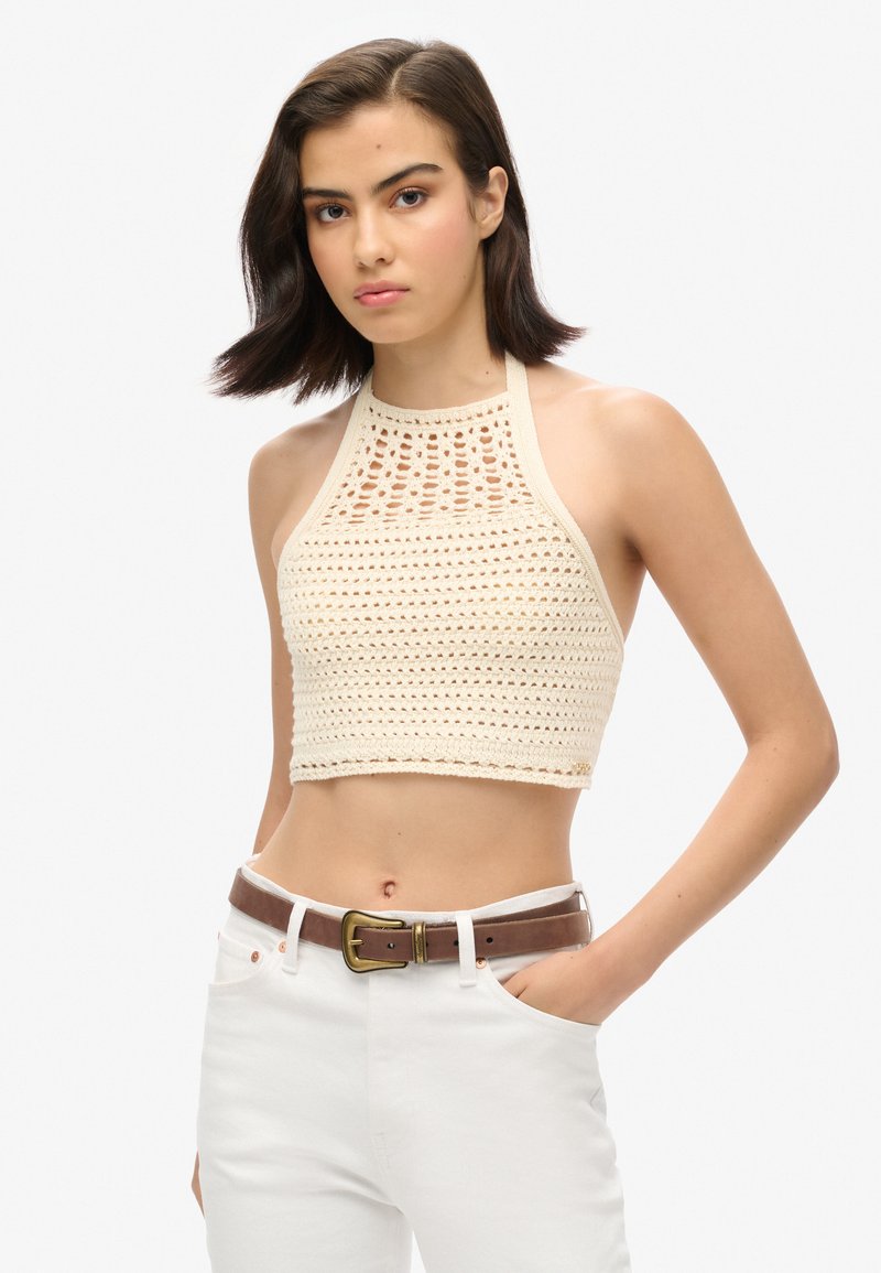Gehaakte haltertop in lichtbeige met een open breipatroon, beschikt over een hoge halslijn en een taille-lange ontwerp, gecombineerd met hoge taille witte jeans.