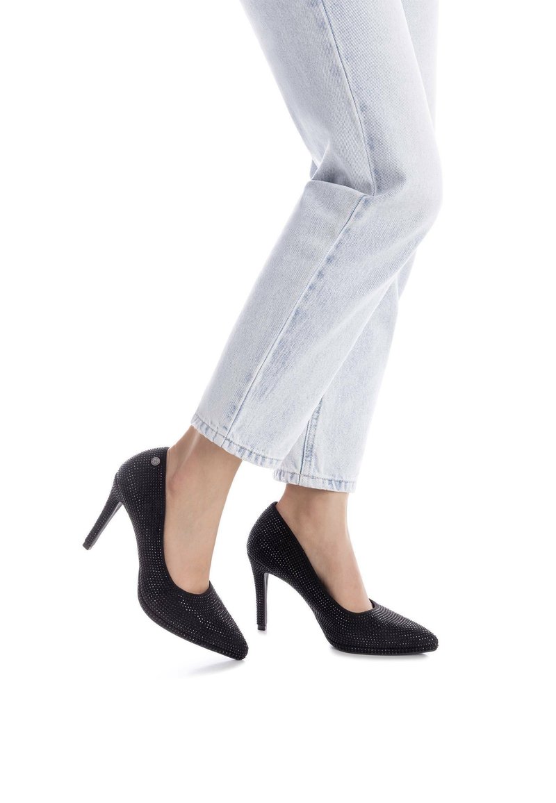 Zapatos de tacón alto negros con punta afilada, adornados con pailletes y una textura suave, llevados con unos vaqueros cortos de color azul claro.