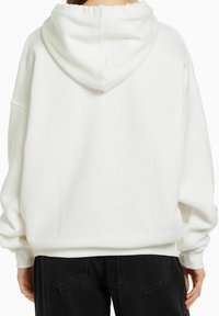 Vit hoodie med justerbar snodd i huvan, oversize passform, ribbad mudd och nederkant, bärs med svarta byxor. Slät tygtextur.