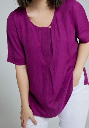 Kobieta ubrana w fioletową bluzkę z krótkim rękawem, mającą pojedynczy guzik przy dekolcie, oraz białe spodnie, z ręką spoczywającą w kieszeni i na udzie.