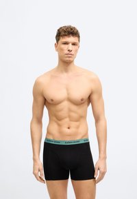 Άνδρας χωρίς πουκάμισο με κοντά σγουρά μαλλιά, φορώντας μαύρα κοντά εσώρουχα με πράσινη μέση που γράφει «BJÖRN BORG», σε απλό φόντο.