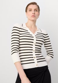 Cardigan blanc à côtes avec des rayures noires, col en V et manches courtes, présentant des boutons dorés sur le devant pour la fermeture.