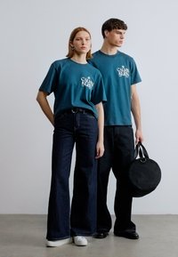 Δύο μοντέλα φορούν τιρκουάζ βαμβακερά t-shirts με λογότυπο, σε συνδυασμό με σκουρόχρωμα τζιν παντελόνια με φούστα. Το αρσενικό μοντέλο κρατά μια στρογγυλή μαύρη τσάντα.