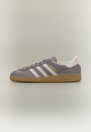 Χαμηλού προφίλ αθλητικό παπούτσι Adidas München σε γκρι σουέτ με λευκές ρίγες, σόλα από καουτσούκ και χρυσό κείμενο "München" στο πλάι.