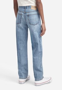 Persona que lleva pantalones vaqueros rectos de color azul claro con bolsillos traseros y etiqueta de Abercrombie, combinados con zapatillas Converse blancas.