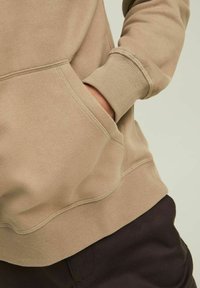 Jasnobrązowa bluza z kieszenią kangurką, ściągaczami przy mankietach i u dołu. Miękka tekstura, minimalistyczny design i luźny krój. Zestawiona z ciemnymi spodniami.
