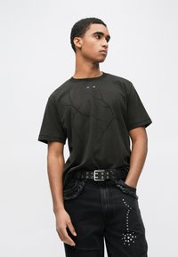 Πράσινο σκούρο κοντομάνικο μπλουζάκι με ανάγλυφο σχέδιο κλαδιού στο μπροστινό μέρος και δύο μικρές οπές κοντά στη λαιμόκοψη, φορεμένο με μαύρο distressed τζιν.