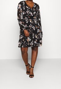 Robe noire à fleurs avec manches longues et col en V, dotée d'un ourlet volanté et d'une taille élastique. Elle présente des motifs floraux colorés.