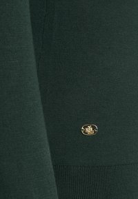 Tissu tricoté vert foncé avec un petit emblème ovale en or présentant les initiales entrelacées « RL » près du bord inférieur.
