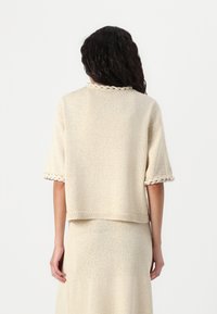 Pull beige à manches courtes avec un tricot texturé, présentant un détail tressé autour du col et des poignets. Design court.