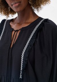 Zwarte jurk met een V-hals, gevlochten striksluiting en witte geborduurde accenten met franjes op de schouders. Zachte stoftextuur.