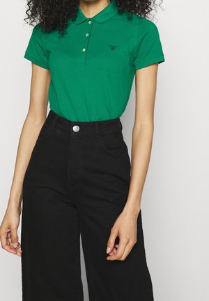 Vrouw met een groen poloshirt met korte mouwen, ingestopt in een zwarte, hoge taille, wijde broek, staand tegen een effen achtergrond.