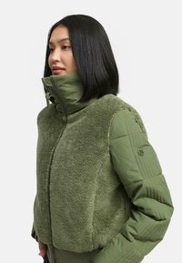 Green jacket featuring a soft, fluffy texture with a contrasting smooth sleeve. It has a high collar and a zip closure, designed for warmth.