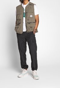 Kaki väst med flera framfickor, bärs över en vit grafisk t-shirt. Kombinationen med svarta cargobyxor och ljusa sneakers med mintgrön detaljer.